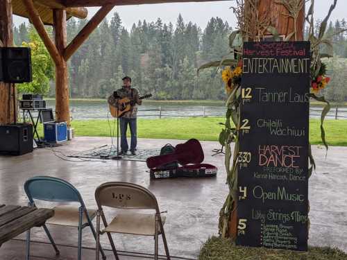 A musician performs on stage at a festival, with a schedule board and scenic backdrop of trees and water.