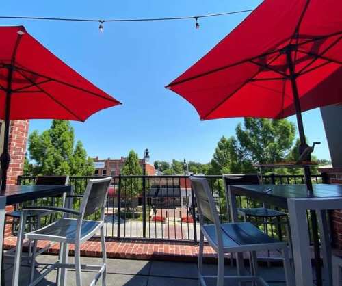 View from a balcony with red umbrellas, overlooking a sunny plaza and trees against a clear blue sky.