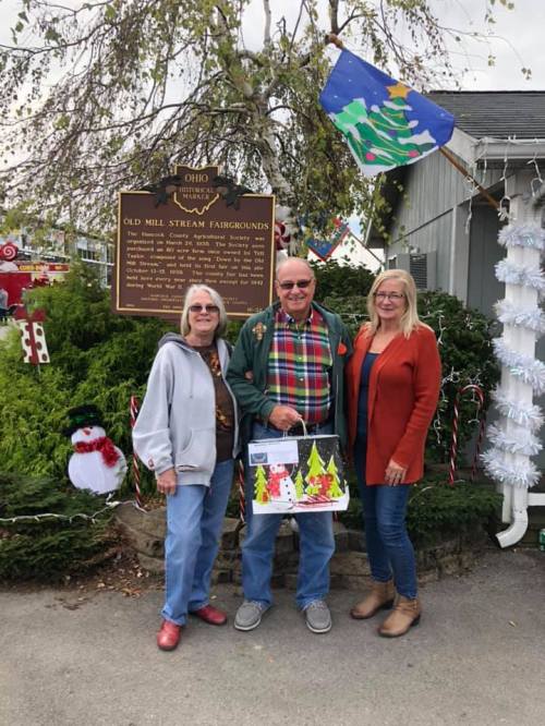 Three people stand in front of a historical sign at a festive outdoor event, surrounded by holiday decorations.