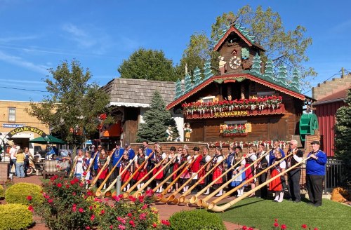 A group of musicians in traditional attire play long horns in front of a decorated wooden building at a festival.