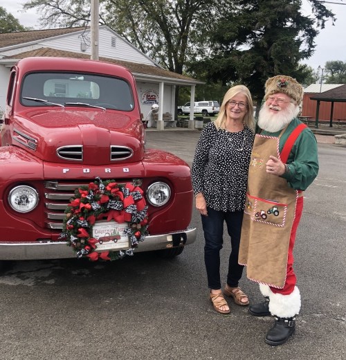 A woman and a man in festive attire stand beside a vintage red Ford truck decorated for the holidays.