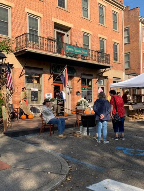 A quaint storefront with pumpkins outside, people chatting, and festive decorations in a charming village setting.