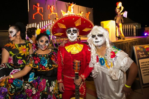 A group of people in colorful costumes and face paint celebrate Día de Muertos at a festive outdoor event.