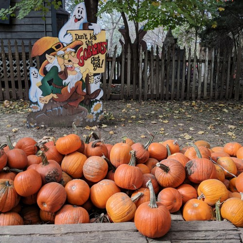 A whimsical sign warns "Don't let the goblins get ya!" near a pile of pumpkins in a fall setting.