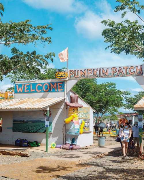 A colorful pumpkin patch entrance with a welcoming sign and a large cartoon character in a hat, surrounded by trees.