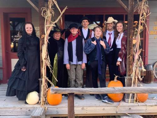 A group of people in costumes poses on a porch decorated with pumpkins and autumn decor.