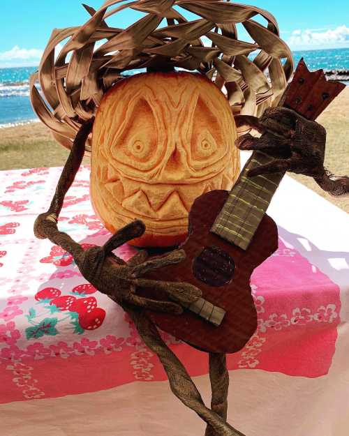 A carved pumpkin with a playful face, wearing a straw hat, holds a small guitar on a colorful tablecloth by the beach.