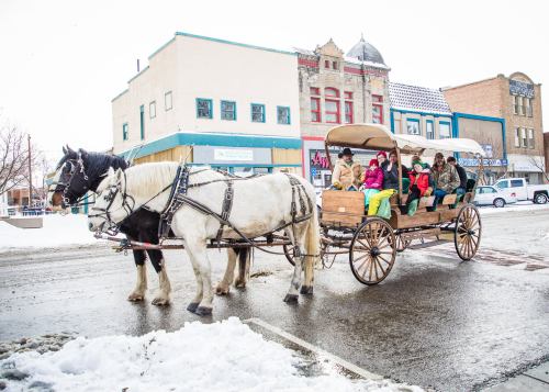 A horse-drawn carriage with passengers rides through a snowy street, surrounded by buildings and parked cars.