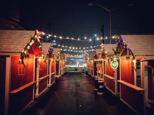 Festive market stalls adorned with colorful lights, set against a dark, rainy night.
