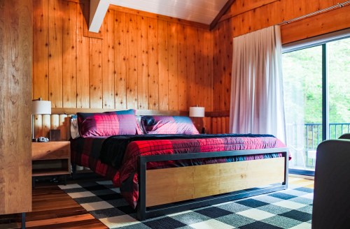 Cozy bedroom with wooden walls, a plaid bedspread, and large windows overlooking greenery.