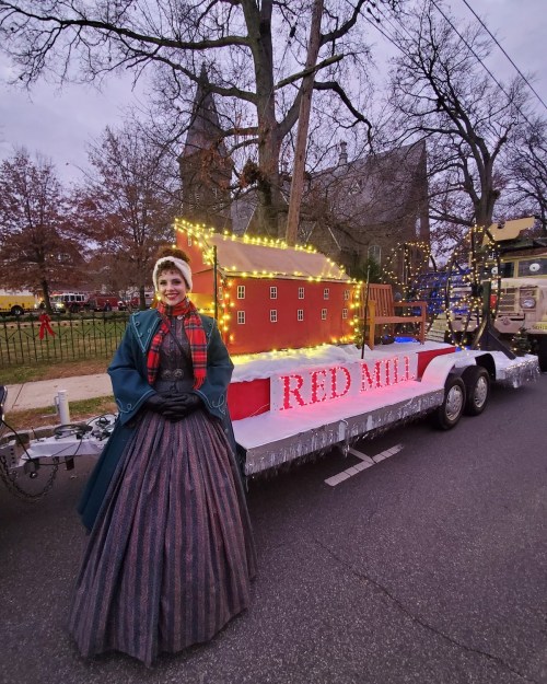 A woman in a vintage dress stands beside a festive parade float decorated with lights and a red mill backdrop.