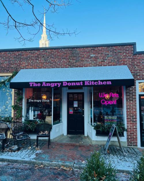 Exterior of The Angry Donut Kitchen with a sign saying "We Are Open!" and a church steeple visible in the background.