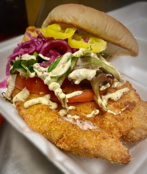 A crispy fish sandwich topped with lettuce, tomatoes, pickled onions, and yellow peppers, served in a soft bun.