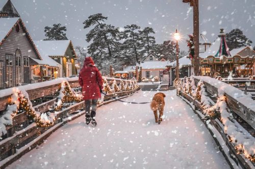 A person in a red coat walks a dog on a snowy path, surrounded by festive decorations and winter scenery.