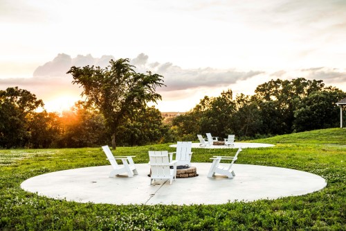 A serene outdoor setting with white chairs arranged around a fire pit, surrounded by green grass and trees at sunset.