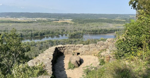 A scenic view from a stone overlook, featuring a river winding through lush green hills and valleys under a clear sky.