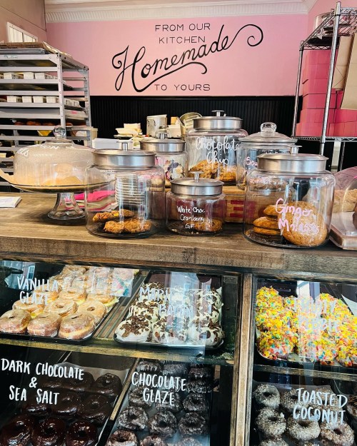 A display of various baked goods in glass jars and trays, with a pink wall that reads "From Our Kitchen Homemade to Yours."