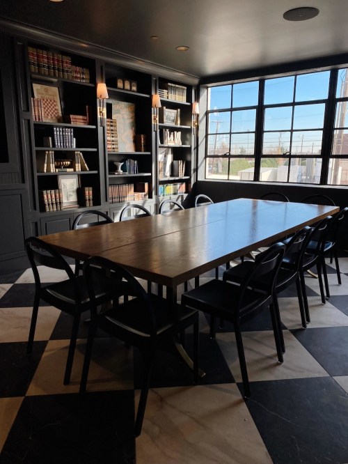 A modern dining room with a large wooden table surrounded by black chairs, featuring bookshelves and a large window.