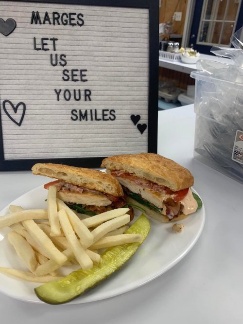 A sandwich with chicken, lettuce, tomato, and sauce, served with fries and a pickle, next to a sign that says "Let us see your smiles."