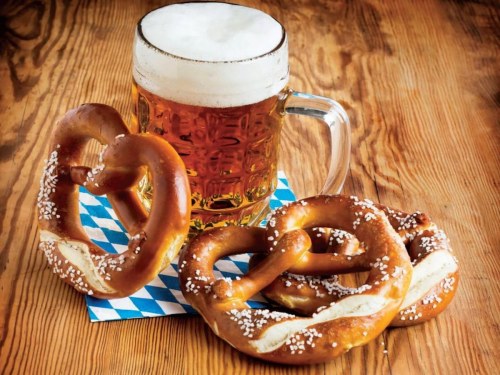 A frothy beer mug next to three soft pretzels on a blue and white checkered napkin, set on a wooden table.