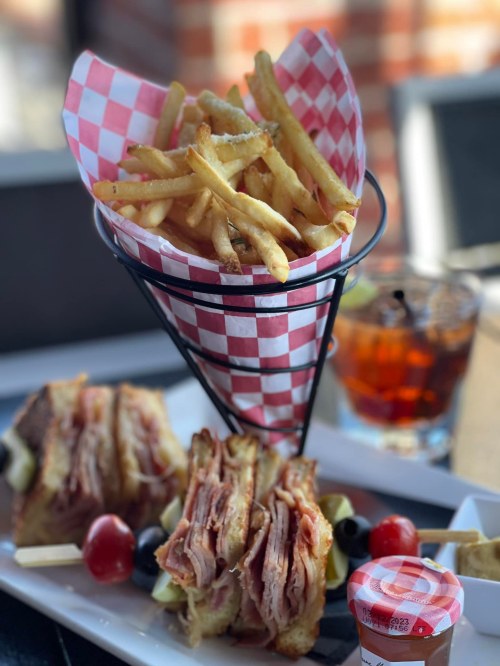 A plate featuring a stacked sandwich, fries in a cone, and a drink, garnished with cherry tomatoes and olives.