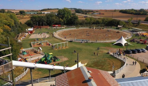 Aerial view of a farm with a circular dirt area, playgrounds, and people enjoying outdoor activities on a sunny day.