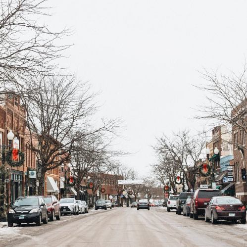 A snowy street lined with shops and decorated with holiday wreaths on bare trees.