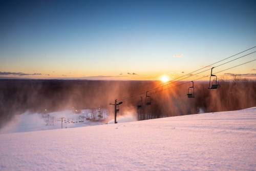 A snowy landscape at sunset, featuring ski lift chairs and mist rising from the slopes.