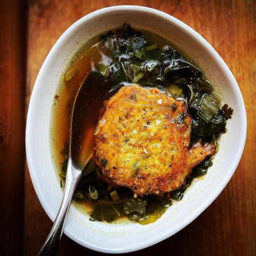 A bowl of soup with greens and a golden-brown fritter, garnished with herbs, served with a spoon.