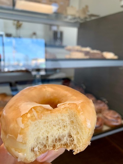 A hand holding a partially eaten glazed donut in front of a display case with various pastries.