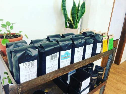 A wooden shelf displaying various bags of Folklore coffee, with plants in the background and coffee cups on the side.