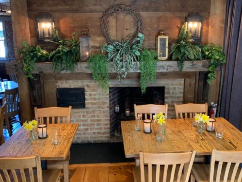 Cozy restaurant interior with wooden tables, yellow flowers, and a decorated fireplace surrounded by greenery.