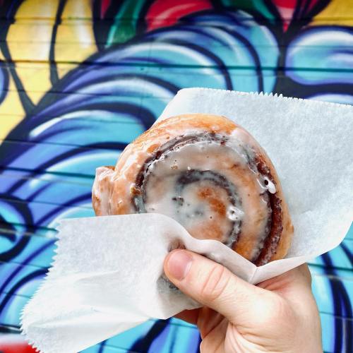 A hand holds a glazed cinnamon roll in front of a colorful mural.