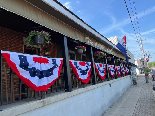 A building's exterior decorated with red, white, and blue bunting, flowers in pots, and flags along the sidewalk.