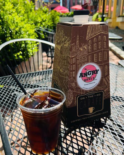 A carton of iced coffee next to a clear cup of iced coffee on a black outdoor table in a sunny setting.