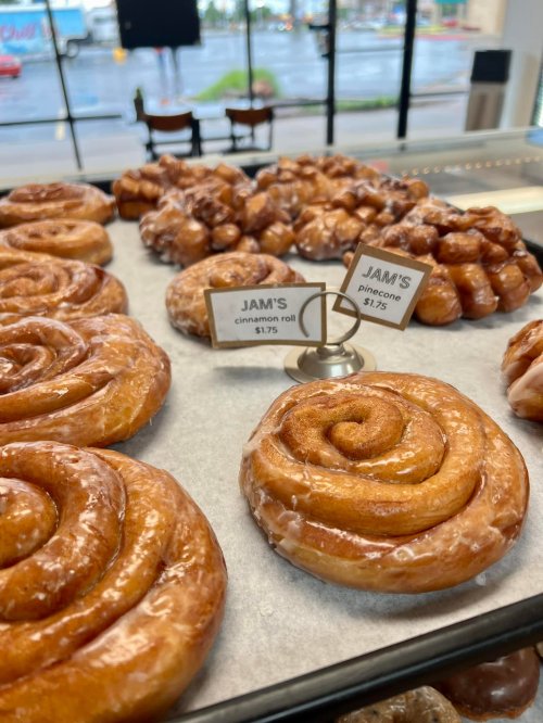 A display of glazed cinnamon rolls and pinecone pastries with price tags in a bakery setting.