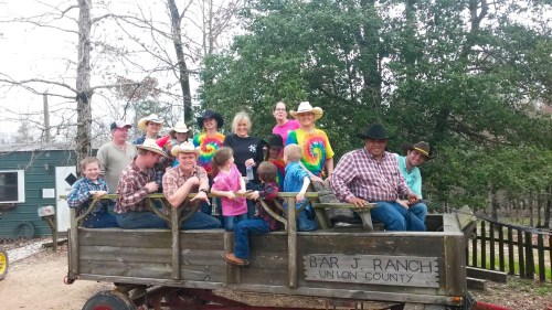 A group of people in cowboy hats and colorful shirts poses on a wagon at Bar J Ranch, surrounded by trees.