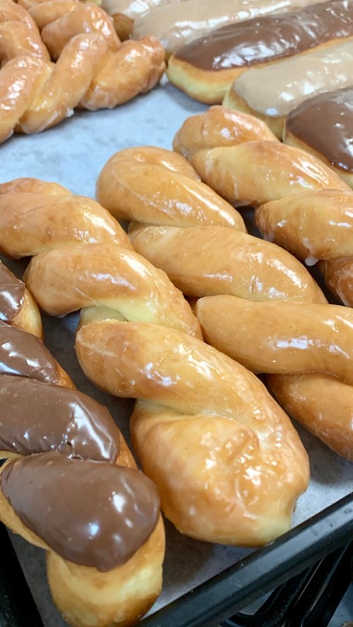 A close-up of glazed twist donuts, some topped with chocolate icing, arranged on a baking tray.