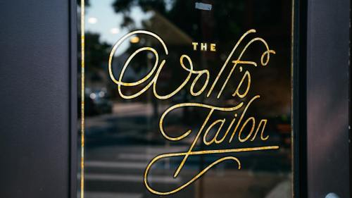 A close-up of a storefront window featuring the elegant sign "The Wolf's Tailor" in gold lettering.