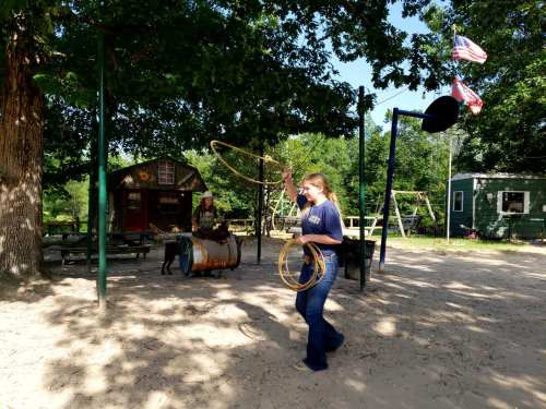 A person swings a lasso in a playground area with trees, a basketball hoop, and a building in the background.
