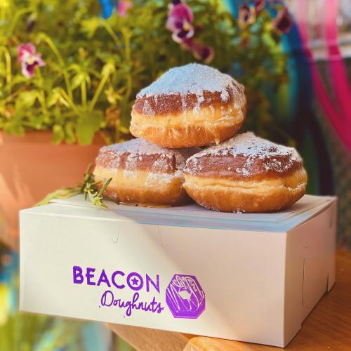 Three powdered sugar doughnuts stacked on a white box with "BEACON Doughnuts" logo, surrounded by colorful flowers.