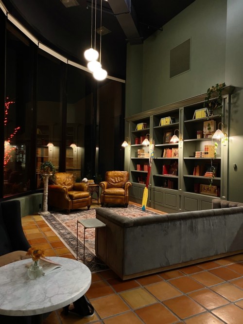 Cozy lounge with two leather chairs, a bookshelf, and warm lighting, featuring a marble table and decorative plants.