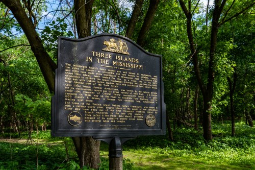 Historical marker titled "Three Islands in the Mississippi" surrounded by lush green trees in a natural setting.