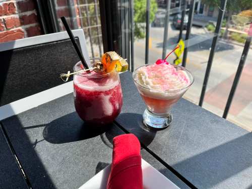Two colorful cocktails on a table: one with a garnish and the other topped with whipped cream and a cherry.