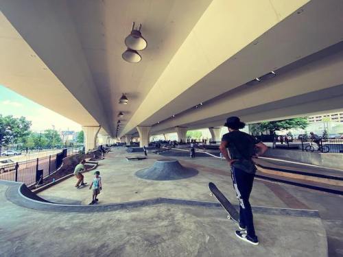A skateboarder watches others at a spacious skatepark under a large concrete structure on a sunny day.