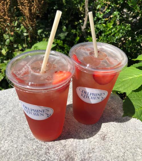 Two clear cups filled with iced tea and strawberries, each with a straw, sitting on a stone surface surrounded by greenery.