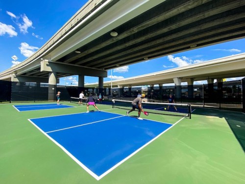 People playing pickleball on blue courts under a highway, with clear skies and clouds in the background.