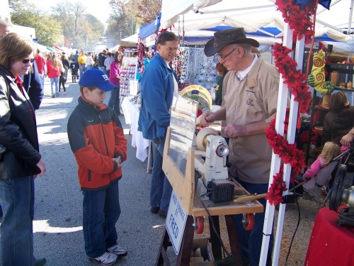 A vendor demonstrates woodturning at a market, while a child watches intently with adults nearby.