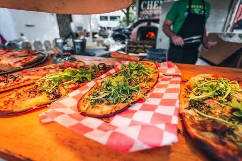 A variety of flatbreads topped with greens and herbs on a wooden table, with a pizza oven in the background.