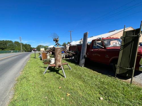 A roadside scene featuring a vintage red truck, various items for sale, and a unique metal sculpture on the grass.